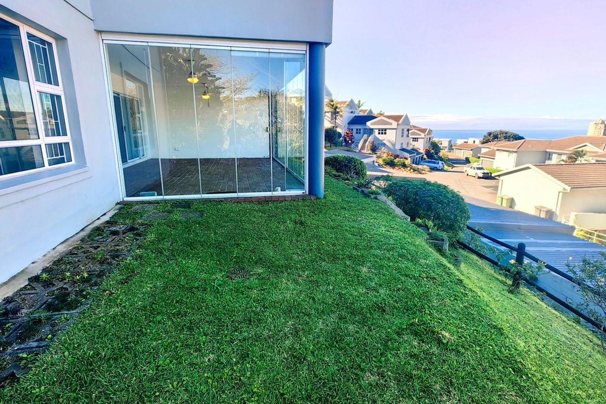 Rear view of the apartment featuring a glass-enclosed patio, upper balcony, and a well-maintained sloped lawn overlooking the neighborhood.