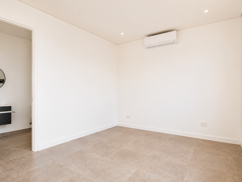 A bright, modern, and empty bedroom featuring neutral tiled flooring, a wall-mounted air conditioning unit, and direct access to an en-suite bathroom.