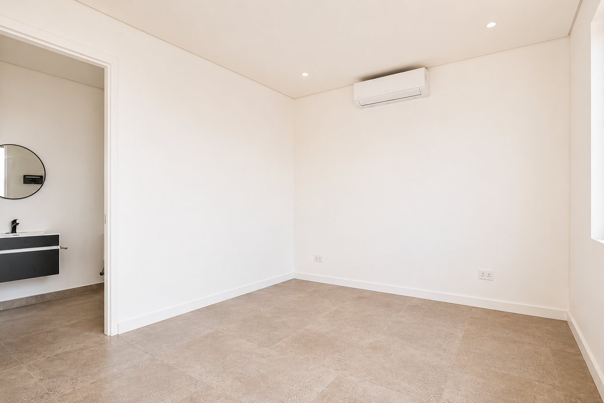 A bright, modern, and empty bedroom featuring neutral tiled flooring, a wall-mounted air conditioning unit, and direct access to an en-suite bathroom.