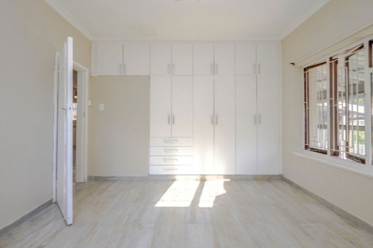 A bright, empty bedroom featuring extensive white built-in cupboards, light-toned laminate flooring, and windows fitted with security bars.