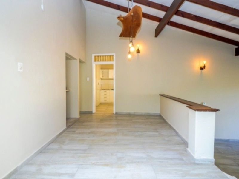 Spacious living area featuring vaulted ceilings with exposed timber beams, tiled flooring, and a half-height partition wall, leading through to a kitchen area.