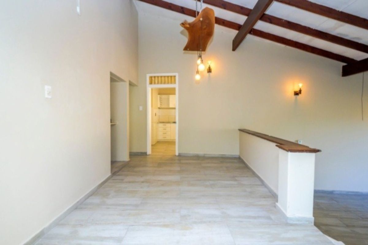 Spacious living area featuring vaulted ceilings with exposed timber beams, tiled flooring, and a half-height partition wall, leading through to a kitchen area.