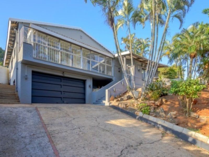 Front elevation of a multi-level house featuring a double garage, concrete driveway, and an enclosed upper-level balcony with established garden surroundings.