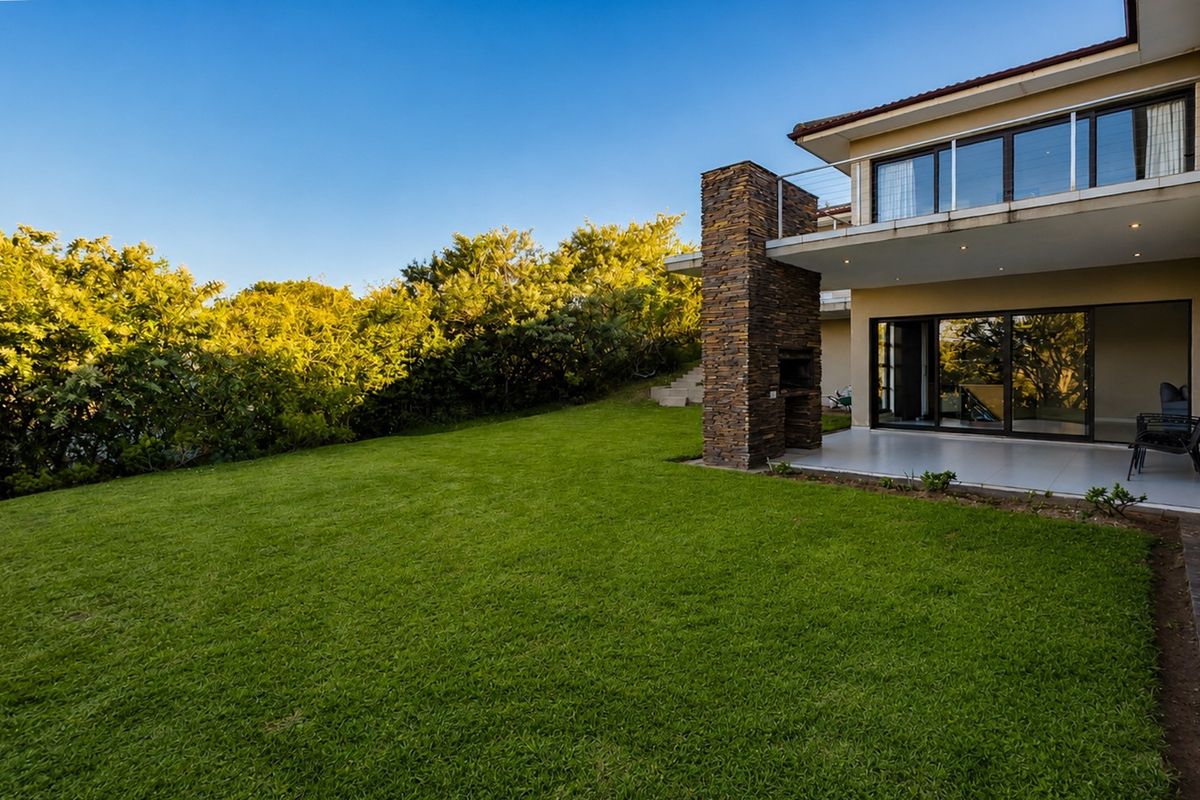 Spacious rear garden featuring a well-maintained lawn, a covered patio with a built-in stone braai, and an upper-level balcony.