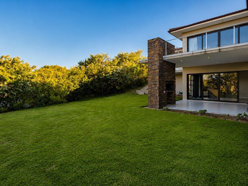 Spacious rear garden featuring a well-maintained lawn, a covered patio with a built-in stone braai, and an upper-level balcony.