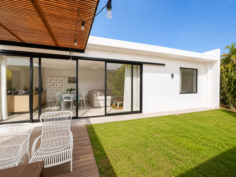 A modern outdoor patio featuring a timber slat pergola, wooden decking, and large sliding glass doors that open into the bright living and kitchen area.
