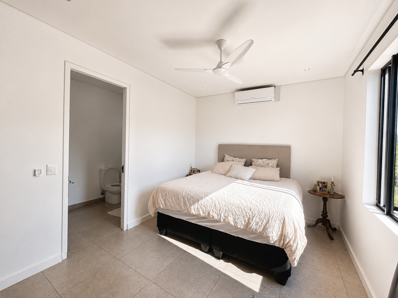A bright and modern bedroom featuring tiled flooring, a ceiling fan, and a wall-mounted air conditioning unit, with direct access to an en-suite bathroom.