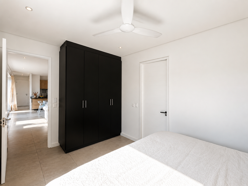 A modern bedroom featuring a sleek black built-in wardrobe, ceiling fan, and neutral tiled flooring, with a view into the adjacent living area.