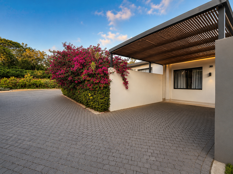 A well-maintained front exterior featuring a paved driveway, a modern timber slat pergola, and established bougainvillea landscaping.