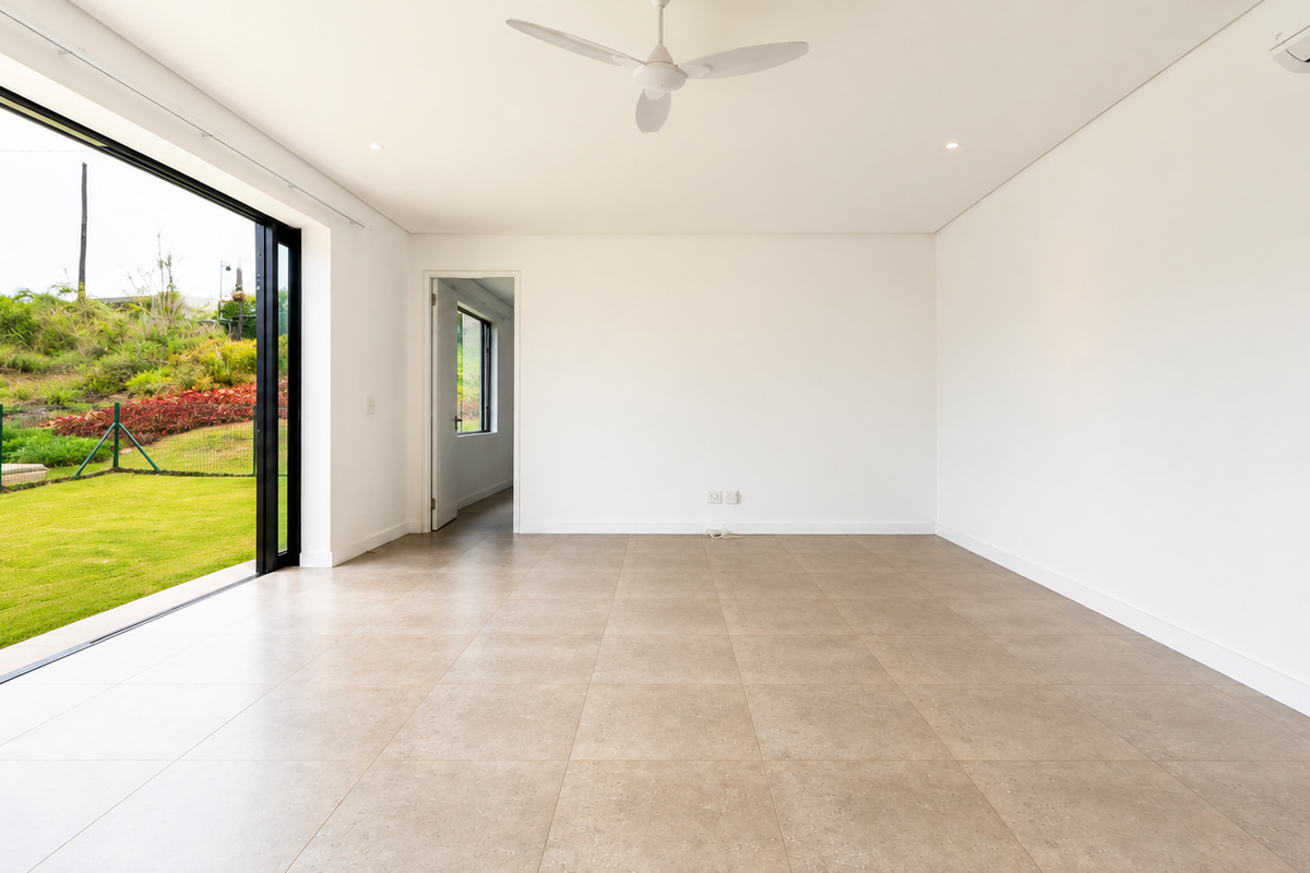 A bright, empty living room featuring large sliding glass doors that open to a garden, tiled floors, a ceiling fan, and air conditioning.