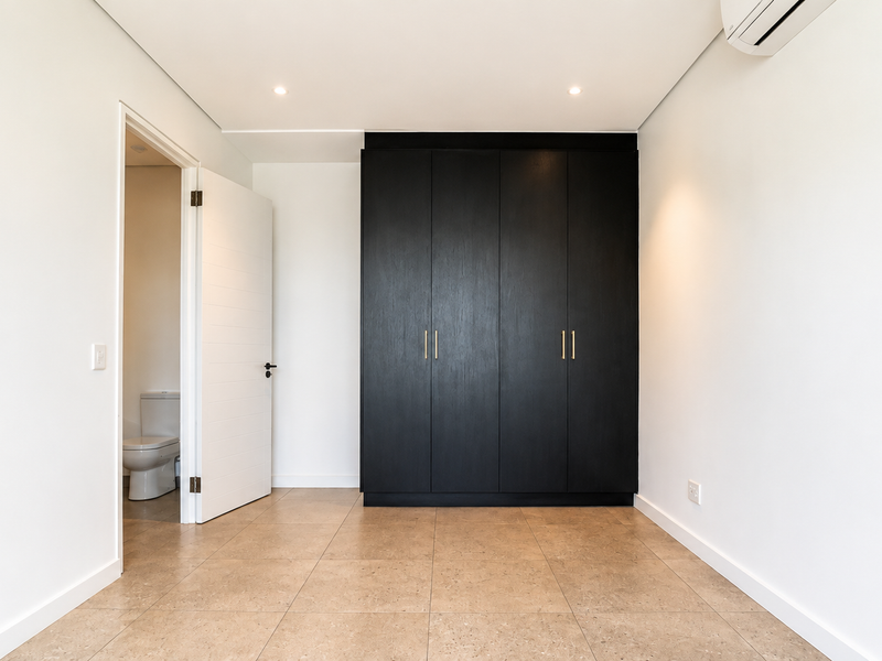 A bright, modern bedroom featuring neutral tiled flooring, a sleek black built-in wardrobe, and an en-suite bathroom.