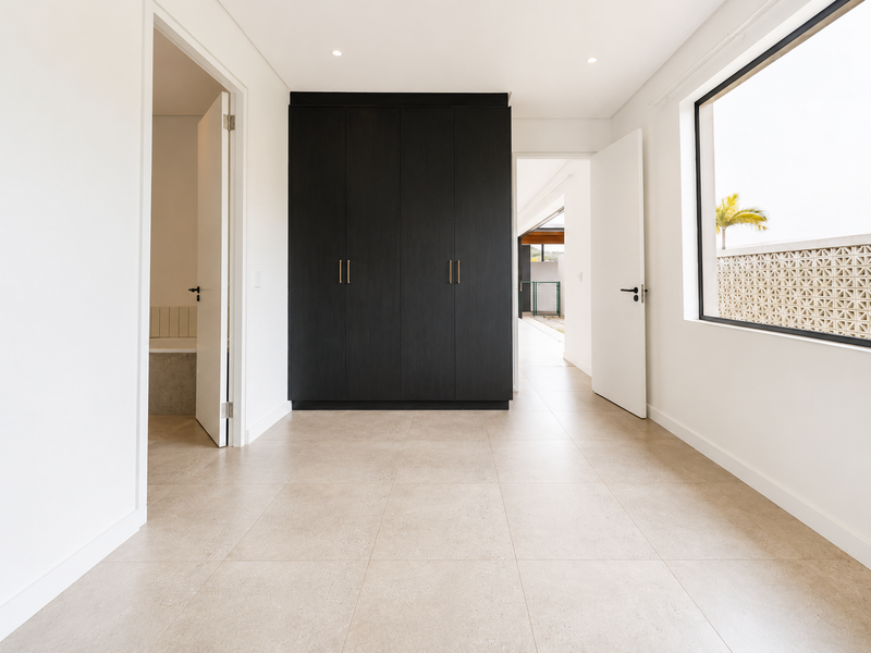 A bright, modern bedroom featuring neutral tiled flooring, dark built-in cupboards, and direct access to an en-suite bathroom.