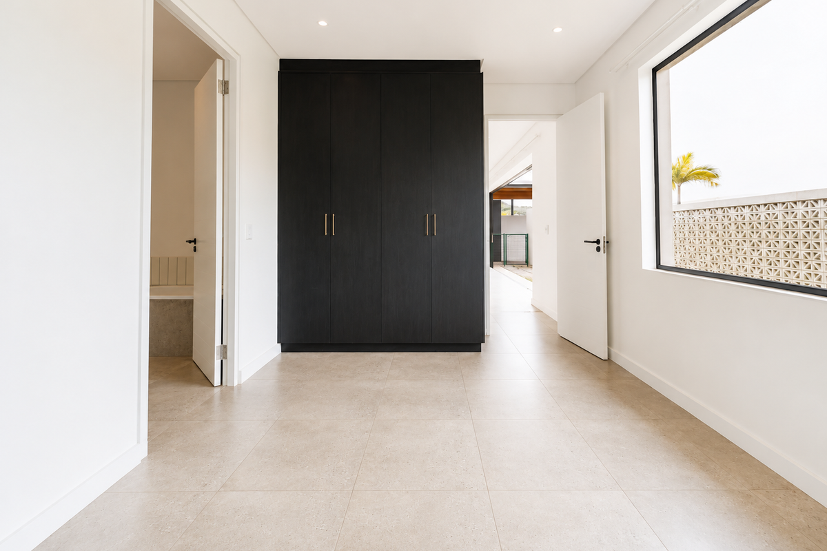 A bright, modern bedroom featuring neutral tiled flooring, dark built-in cupboards, and direct access to an en-suite bathroom.