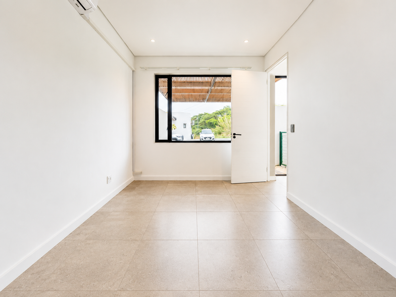 A bright, empty living room featuring neutral tiled flooring, recessed lighting, and a split-system air conditioner, with direct access to an outdoor area.