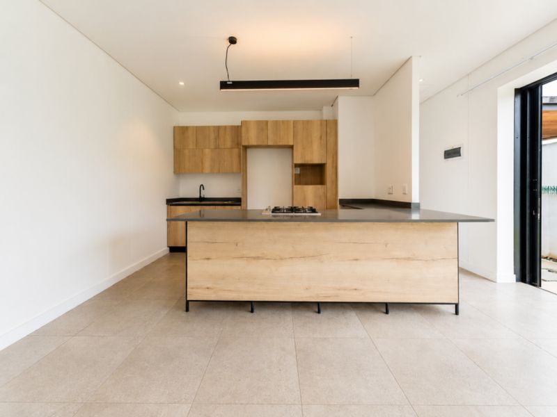 Modern open-plan kitchen featuring light wood cabinetry, a central island with a gas hob, dark countertops, and large format floor tiles.