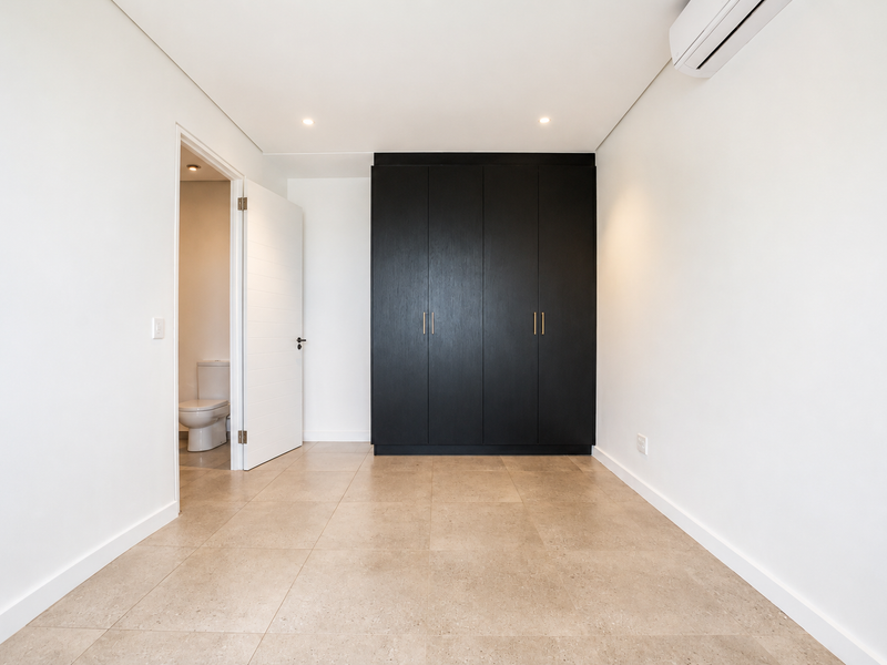 A bright, modern bedroom featuring neutral tiled flooring, a sleek black built-in wardrobe, and a wall-mounted air conditioning unit.