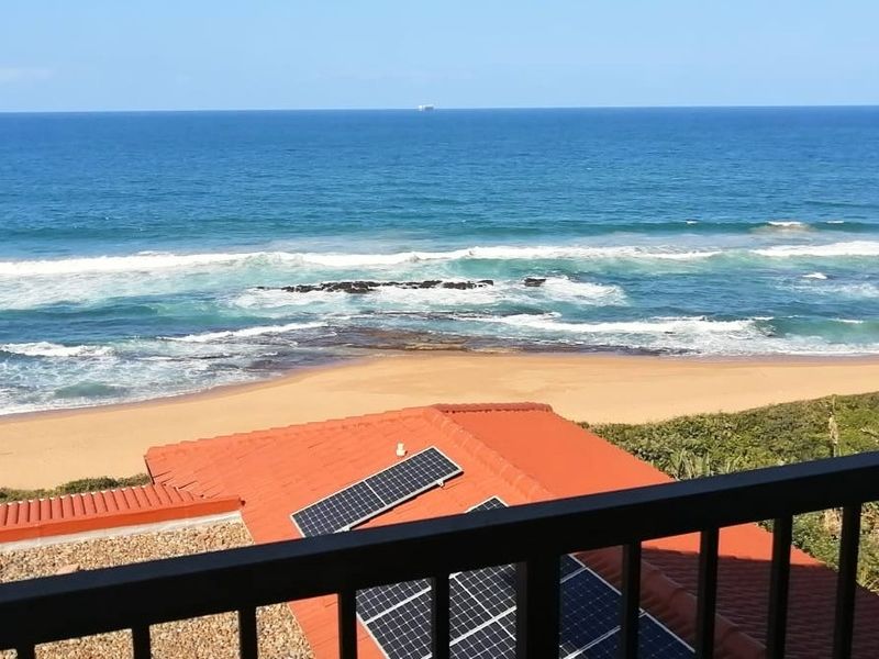 A scenic elevated view overlooking the ocean and sandy beach, featuring a tiled roof with integrated solar panels and a balcony railing in the foreground.