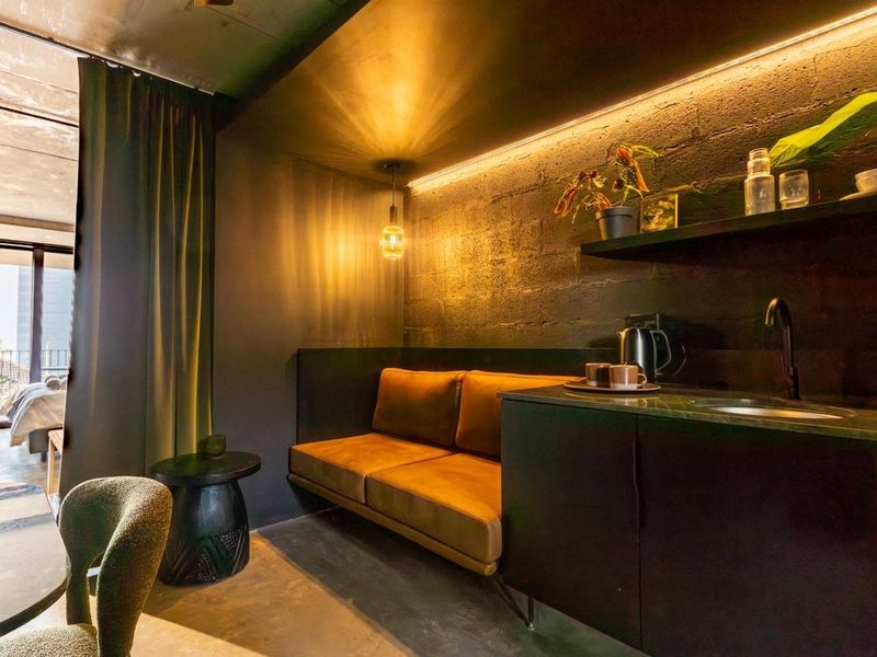 Modern open-plan studio apartment featuring a kitchenette, exposed brick and concrete finishes, warm ambient lighting, and direct access to a private balcony.