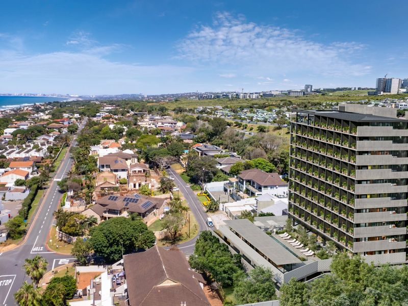 Aerial view of a modern multi-story apartment complex featuring a swimming pool and landscaped grounds, situated in a coastal residential neighborhood.
