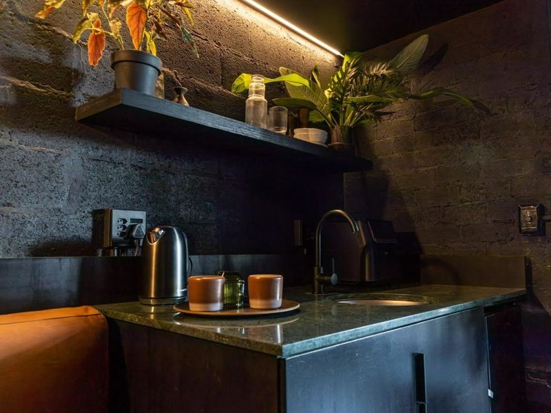 A modern, moody scullery area featuring dark exposed brick walls, a stone countertop with an integrated sink, and sleek built-in cabinetry with accent lighting.