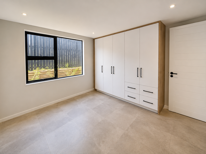 A bright, modern bedroom featuring neutral tiled flooring, recessed downlights, and a large built-in wardrobe with white cabinetry and black hardware.
