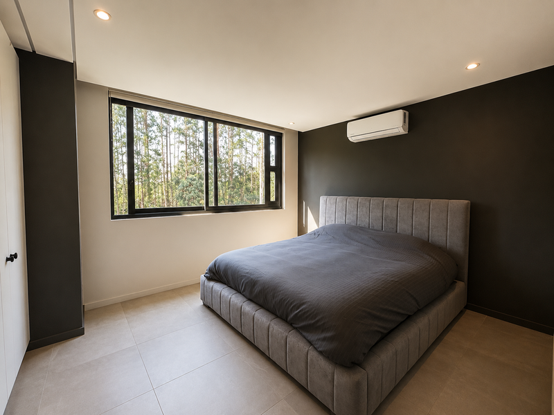 Modern bedroom featuring neutral tiled flooring, dark accent walls, a split-system air conditioner, and large sliding windows with a natural forest view.