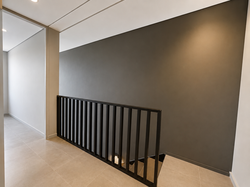 A modern, minimalist hallway featuring neutral tiled flooring, a striking dark grey accent wall, and a sleek black metal staircase balustrade.