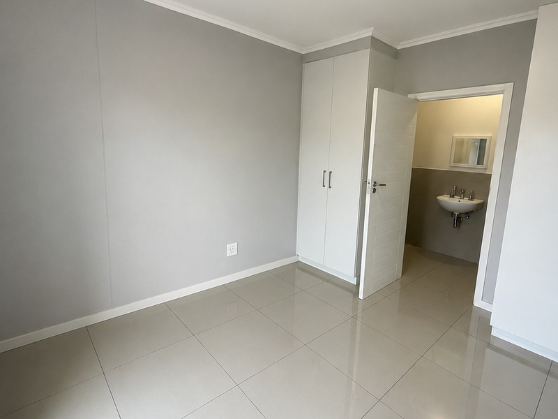A bright, modern bedroom featuring neutral grey walls, large format floor tiles, and ample built-in cupboard space with direct access to an en-suite bathroom.