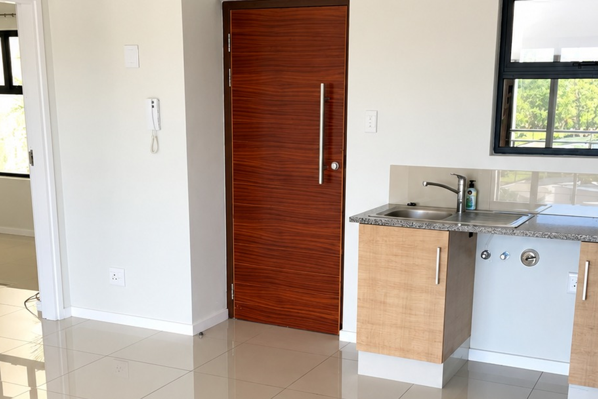 A clean, modern kitchen area featuring light wood cabinetry, granite countertops, a stainless steel sink, and polished floor tiles in an open-plan apartment layout.