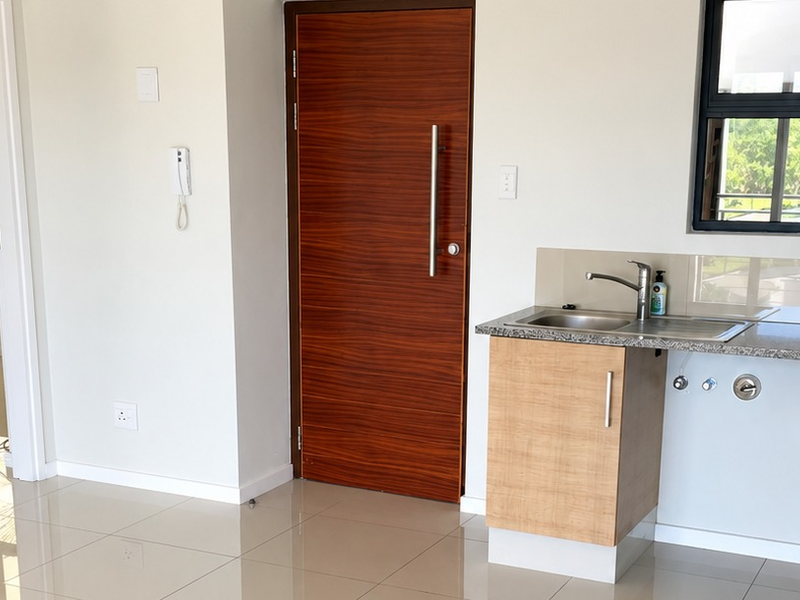 A clean, modern kitchen area featuring light wood cabinetry, granite countertops, a stainless steel sink, and polished floor tiles in an open-plan apartment layout.