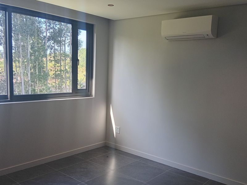 A bright, modern, and empty bedroom featuring tiled flooring, a wall-mounted air conditioning unit, and large windows with a pleasant outdoor view.