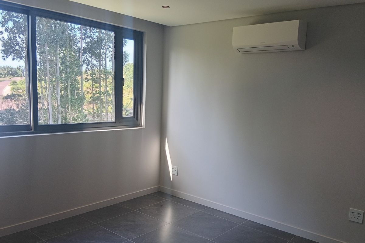 A bright, modern, and empty bedroom featuring tiled flooring, a wall-mounted air conditioning unit, and large windows with a pleasant outdoor view.