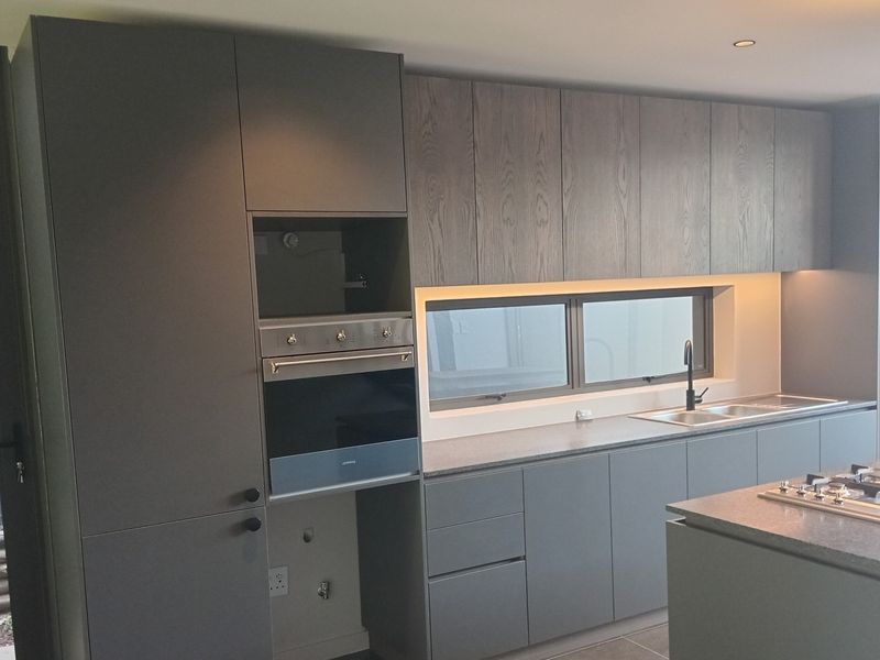 Modern kitchen featuring sleek grey cabinetry, a central island with a gas hob, built-in oven, and integrated stainless steel sink with contemporary finishes.