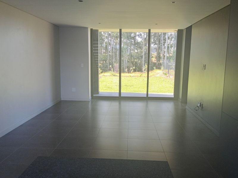 A bright, modern open-plan living area featuring large floor-to-ceiling glass doors, neutral tiled flooring, and recessed lighting, offering a seamless transition to the outdoors.