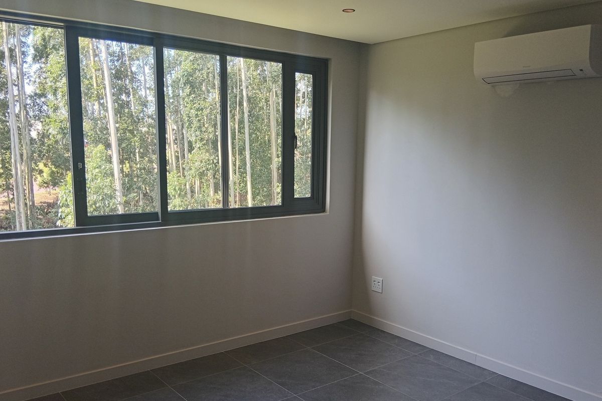 A bright, empty bedroom featuring modern grey floor tiles, a large aluminium sliding window with a forest view, and a wall-mounted split-system air conditioner.