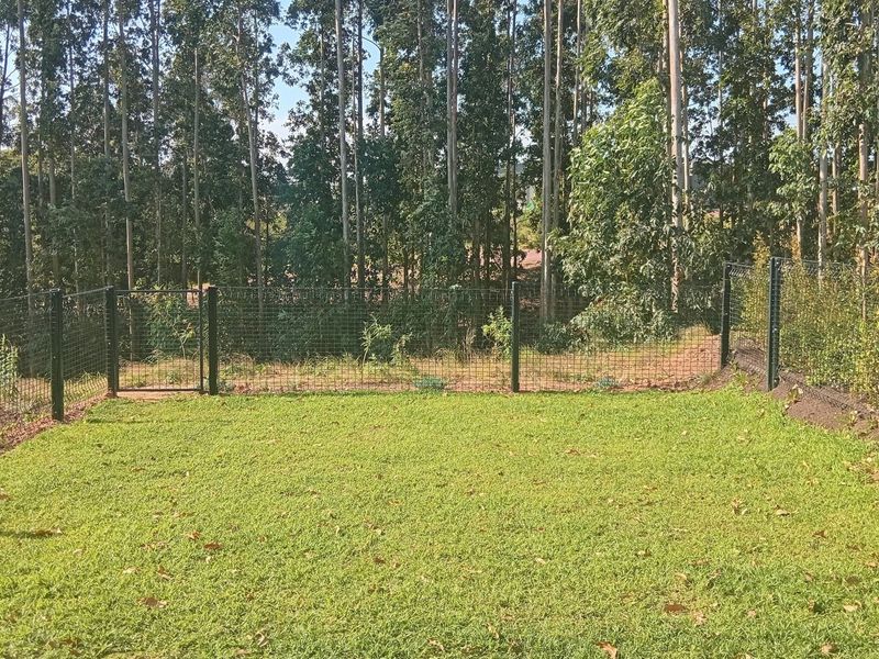 A private backyard garden featuring a well-maintained lawn, enclosed by wire mesh fencing and a metal gate, backing onto a wooded area.