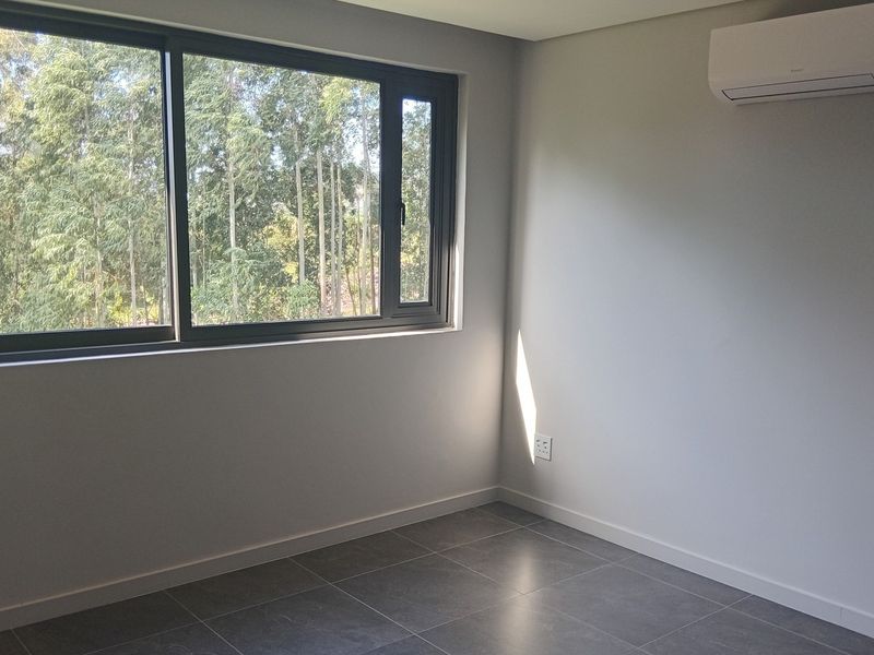 A bright, empty bedroom featuring modern grey floor tiles, a large sliding window with a forest view, and a wall-mounted air conditioning unit.
