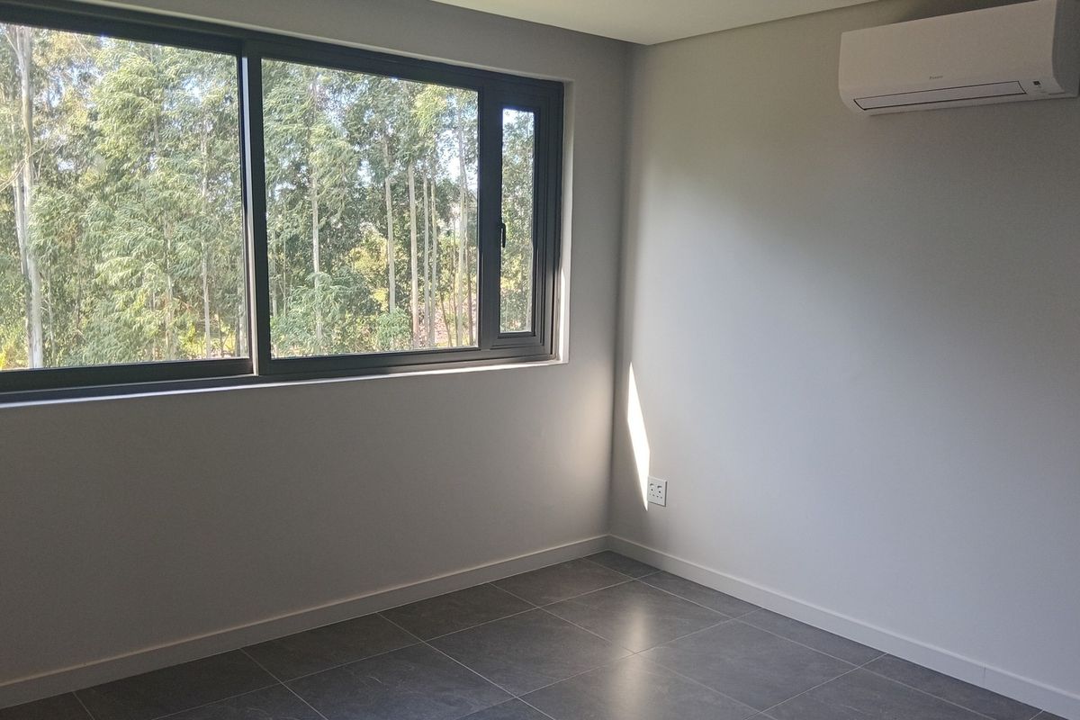 A bright, empty bedroom featuring modern grey floor tiles, a large sliding window with a forest view, and a wall-mounted air conditioning unit.