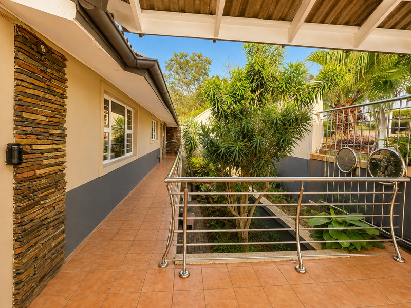 A covered outdoor balcony featuring tiled flooring, a decorative stone-clad wall, and a modern stainless steel balustrade overlooking a landscaped garden area.