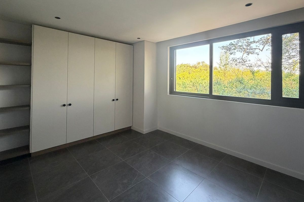 A bright, modern bedroom featuring large format floor tiles, extensive built-in wardrobe storage with integrated shelving, and a large window with garden views.