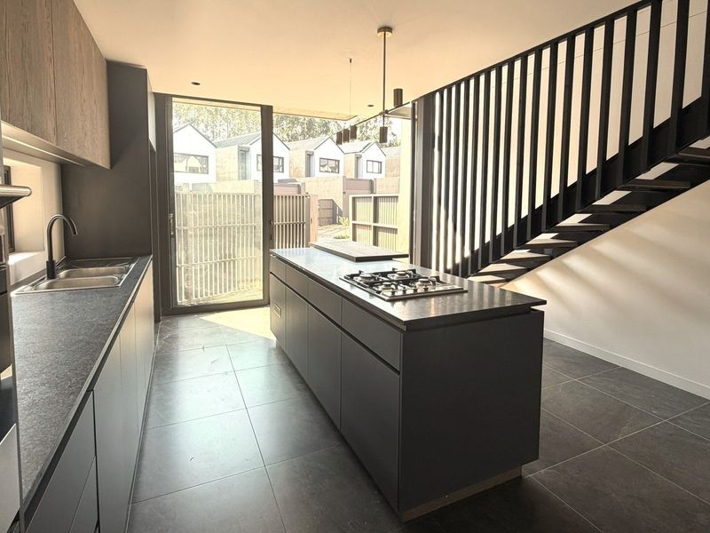 Modern kitchen featuring a central island with gas cooktop, sleek cabinetry, tiled floors, and an adjacent staircase, leading to an outdoor patio area.