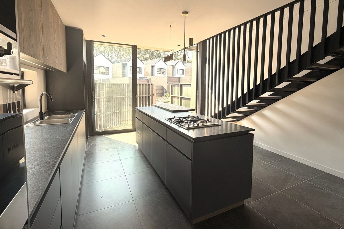 Modern kitchen featuring a central island with gas cooktop, sleek cabinetry, tiled floors, and an adjacent staircase, leading to an outdoor patio area.
