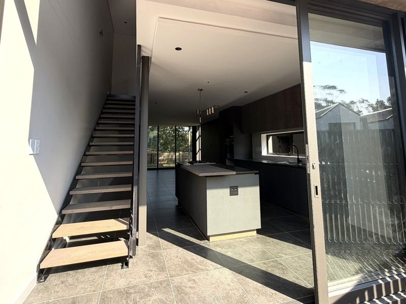 Modern open-plan kitchen and living area featuring a central island, sleek cabinetry, and a contemporary staircase, seamlessly connected to the outdoors via sliding glass doors.