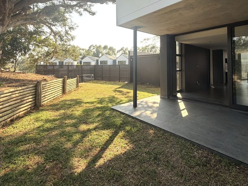 A private rear garden featuring a tiled patio area, timber retaining wall, and mature trees, providing a seamless transition from the indoor living space.