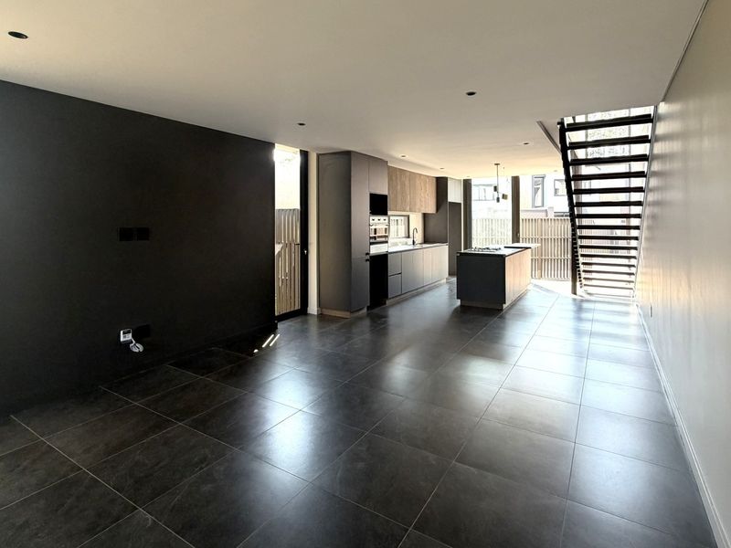 Modern open-plan living and kitchen area featuring dark cabinetry, a central island, sleek tiled flooring, and a contemporary staircase in a bright, spacious layout.