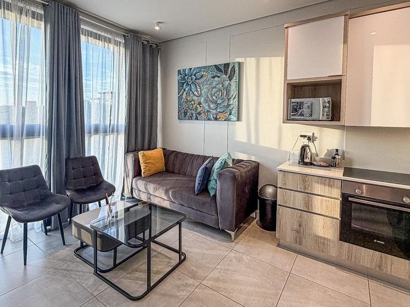 Modern open-plan living area featuring a compact kitchen with built-in appliances, tiled floors, and large windows providing ample natural light.