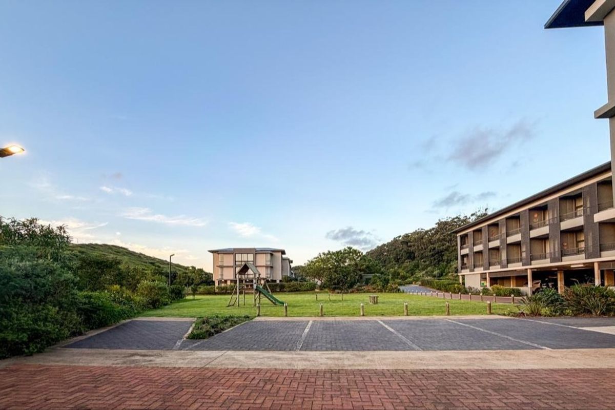 A view of the apartment complex exterior featuring a paved parking area, well-maintained lawn, and surrounding landscaped grounds.
