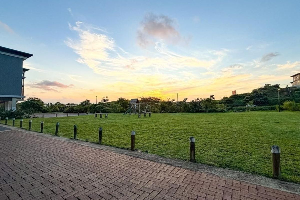 A well-maintained communal garden area featuring a paved walkway, timber bollards, and expansive lawn space at sunset.