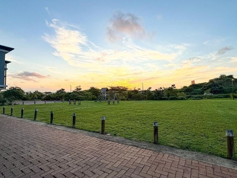 A well-maintained communal garden area featuring a paved walkway, timber bollards, and expansive lawn space at sunset.