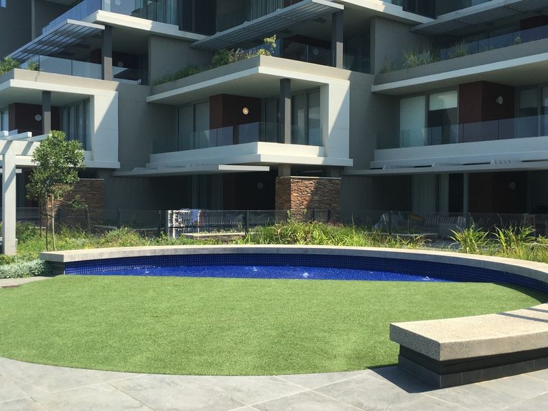A well-maintained communal pool area featuring a circular pool with blue tiling, artificial turf, and modern apartment building architecture in the background.